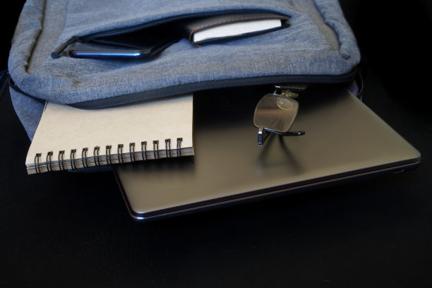 黑色背景上放着一个灰色城市背包，里面有一台笔记本电脑或超级本(ultrabook)、眼镜、一部智能手机和一个记事本。城市旅行者、程序员或学生的属性。自由空间图片下载