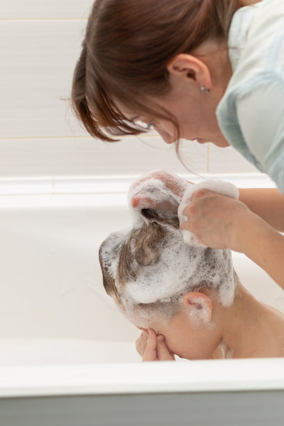 女母亲用洗发水在白色的背景上用手给儿子洗头。男孩眯着眼睛，用手捂住眼睛图片下载