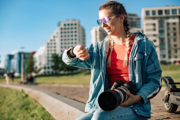 一位年轻的女摄影师正在户外拍摄。女孩坐在草地上，看着智能手表上的时钟并拍照图片下载