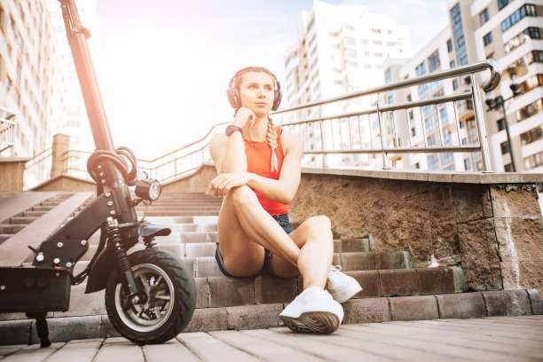 使用电动滑板车的女人的肖像。推滑板车概念户外街道。一个人骑着电动车坐在楼梯上放松。照片与音乐无线耳机图片下载