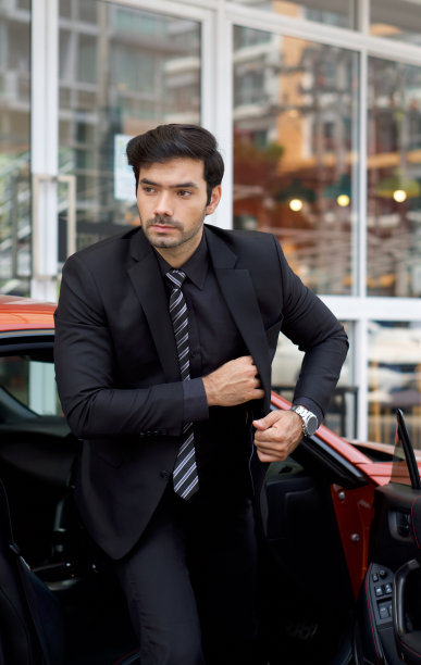 穿着黑色西装的年轻商人从一辆亮闪闪的跑车上走下来，把他的东西放进西装口袋里。图片下载