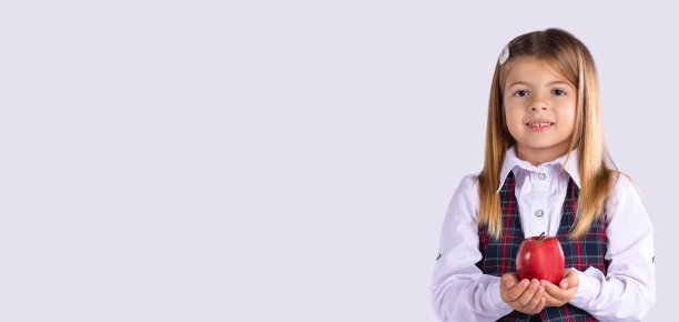 横幅,长格式。小学生的健康零食。一个穿着校服的女孩摆姿势，手里拿着一个红苹果。灰色工作室背景。图片下载