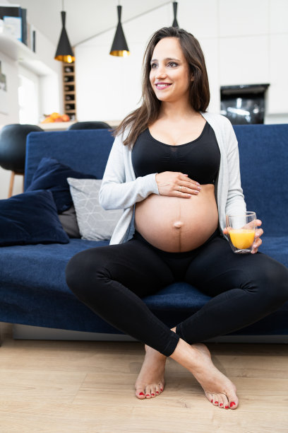 年轻孕妇坐在沙发上的肖像，触摸她的肚子在她的家的客厅。微笑的女性与一杯橙汁在她的手。背景中的厨房图片下载