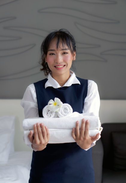 年轻的酒店女服务员微笑着拿着一套带有白花的毛巾。身着蓝色制服的工作人员为酒店客人准备房间。图片下载