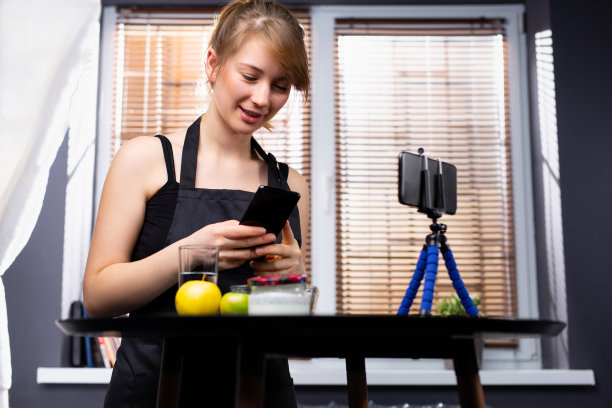 这位金发白种人在她的手机摄像头前用自拍杆上传了一个关于健康饮食的视频博客，并拍下了她烹饪的食物。图片下载