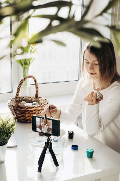 年轻女子博客直播在线研讨会。展示如何装饰复活节彩蛋。三脚架智能手机，篮子鸡蛋，油漆和笔记本电脑。冠状病毒封锁期间的传统节日图片下载