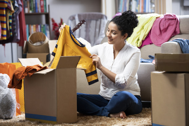 一个年轻女人拿着衣服打开箱子，坐在家里的地板上:库存照片图片下载