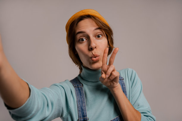 照片的可爱的女性潮人自拍，显示和平和爱的姿态。白人妇女穿着牛仔整体和帽子孤立的灰色背景图片下载