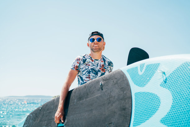 在阳光明媚的中午，一名金发碧眼的冲浪者戴着蓝色太阳镜和帽子，背着冲浪板。暑假期间活跃的人近海概念形象。图片下载