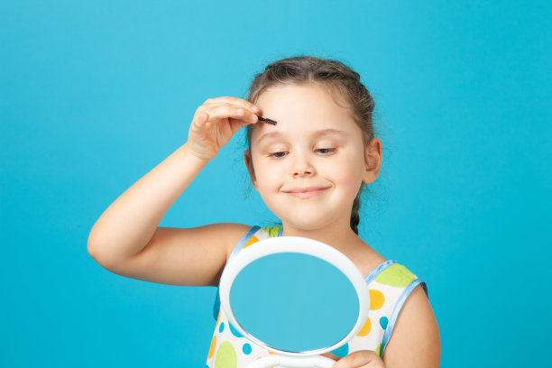 一个穿着白色连衣裙的积极迷人的女孩在镜子前用刷子梳着眉毛，孤立在蓝色的背景上图片下载