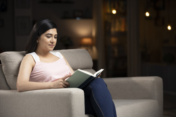 一个年轻女人坐在家里的沙发上看书的照片:-库存照片图片下载