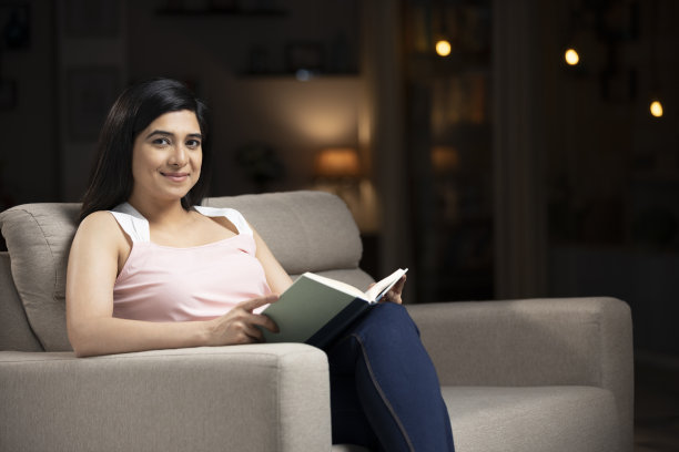 一个年轻女人坐在家里的沙发上看书的照片:-库存照片图片下载