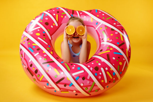 夏天的概念。拿着半个橙子的小女孩躺在一个充气甜甜圈上。画室里黄色背景下的孩子。图片下载