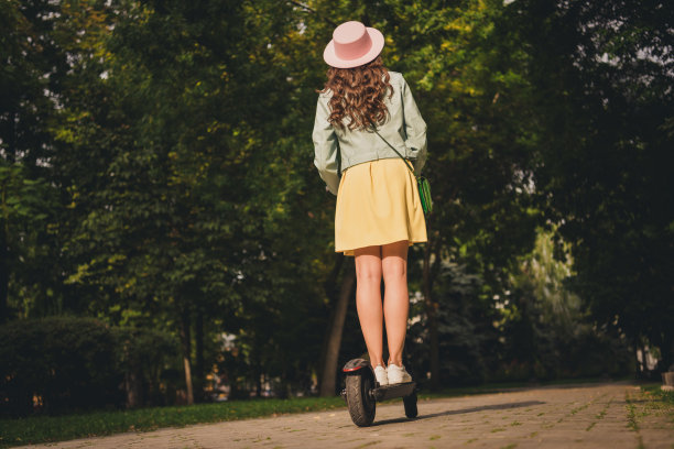 照片苗条的女孩年轻的女士穿绿松石夹克，头戴手提包驾驶快速滑板车城外的城市公园图片下载
