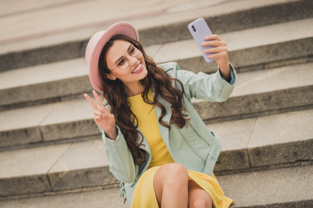 肖像迷人的女孩坐在楼梯上，使自拍手指显示v字有好心情，在户外自由时间图片下载