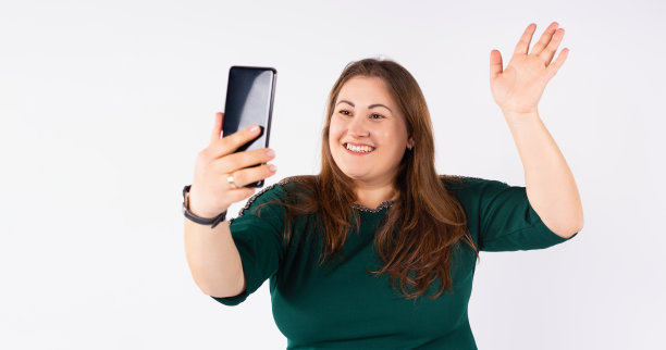 快乐丰满的女人微笑着，用她的智能手机拍了一张自拍，挥手摆姿势。工作室白色背景。横幅。图片下载