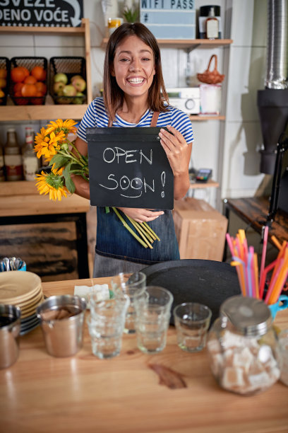快乐的年轻女性拿着向日葵和写着“即将开放”的牌子，为她的新事业兴奋不已。幸福，快乐，兴奋，开始，开始，事业图片下载