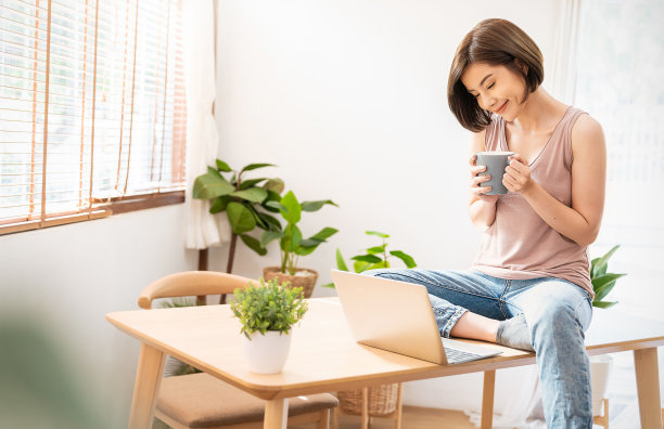 年轻美丽的亚洲女人的肖像放松坐在书桌上使用电脑笔记本电脑。微笑快乐的亚洲女孩在家喝咖啡。自由小企业主中小企业，在家工作的概念。图片下载