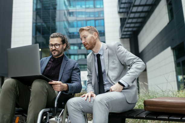 一个坐着轮椅的年轻商人和他的同事坐在外面。库存图片图片下载