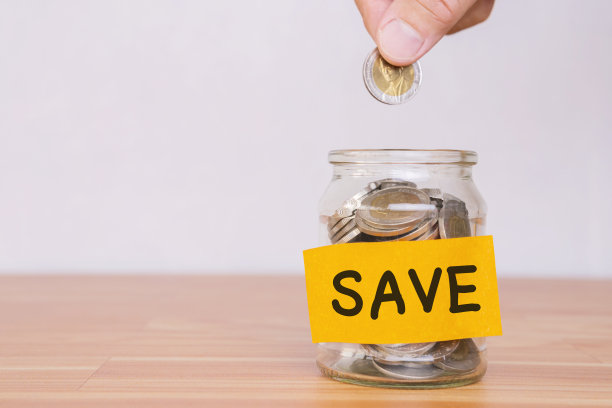 亚洲女人手里正把一枚硬币放在一个玻璃瓶里，一堆硬币放在一张棕色的木桌上素材图片