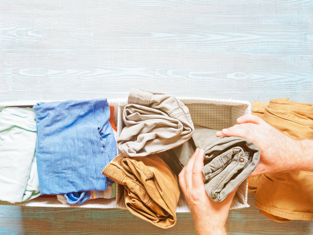 男士的手将卷起的裤子(裤子、牛仔裤)从箱子里拿出来或放进箱子里。整理男士衣柜的概念。附近的抽屉里还有其他衣服。木制背景。图片下载