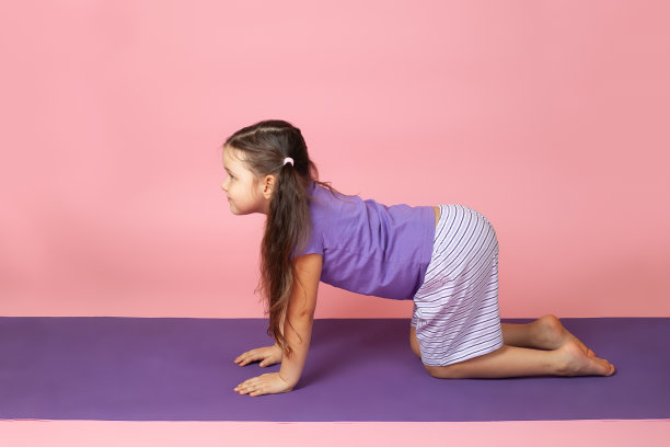 一个5岁的小女孩在紫色的瑜伽垫上做猫式或马尔贾利亚式，在粉红色的背景上孤立地做瑜伽图片下载