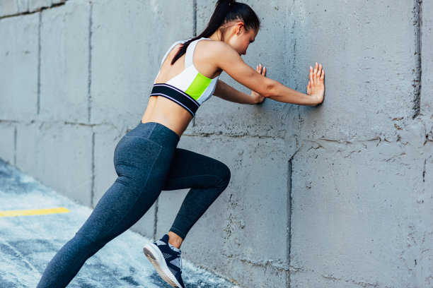 后方的健身年轻女子做伸展运动在混凝土灰色的墙壁。女运动员在户外晨练。图片下载