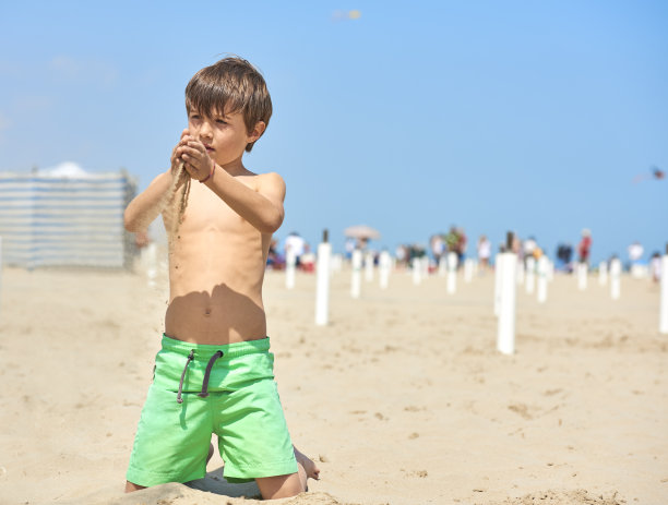阳光灿烂的日子里，那个穿着游泳衣的孩子在海滩上玩沙子。背景是人们在海边散步的海滩。图片下载