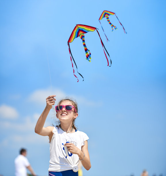 穿t恤的小女孩拿着五颜六色的风筝在海边的沙滩上玩。背景中有许多人在海滩上散步。图片下载