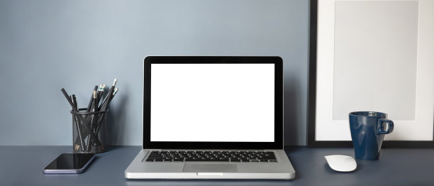 电脑笔记本前视图，白色屏幕，智能手机，咖啡杯，笔筒和空框架在深蓝色的桌子上。时尚的工作场所。图片下载