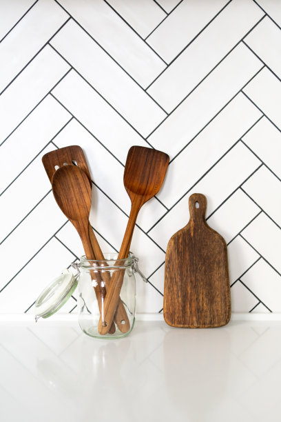 国内的厨房用具。玻璃罐中的木制铲和切菜板，背景为白色几何瓷砖。图片下载