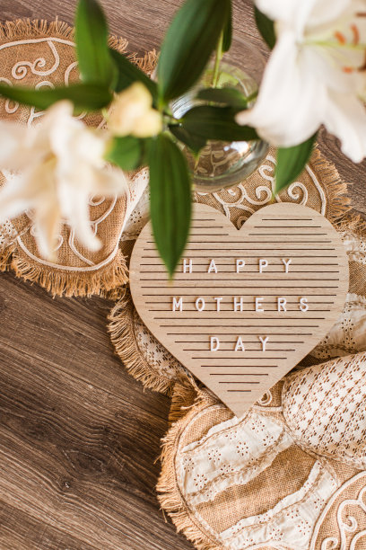 “母亲节快乐”的字样写在一个心形的广告牌上，周围是白色的兰花和复活节百合，背景是粗麻布和花边图片下载