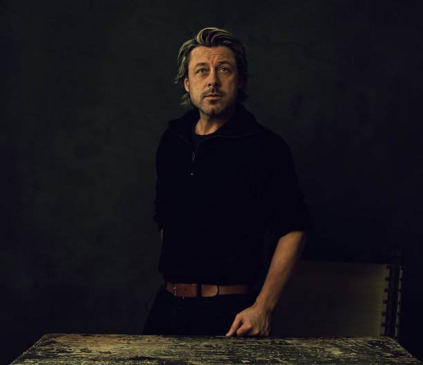 一个严肃的留着胡茬胡子的金发男人穿着黑色羊毛衫和牛仔裤站在一堵黑墙前的老式桌子后面。图片下载