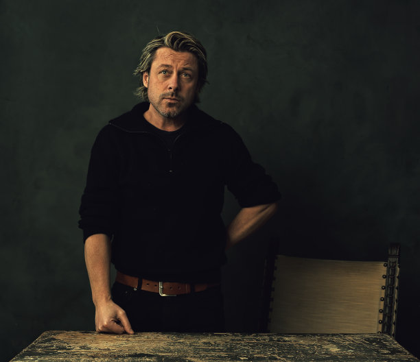 一个严肃的留着胡茬胡子的金发男人穿着黑色羊毛衫和牛仔裤站在一堵黑墙前的老式桌子后面。图片下载