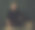 一个严肃的留着胡茬胡子的金发男人穿着黑色羊毛衫和牛仔裤站在一堵黑墙前的老式桌子后面。图片下载