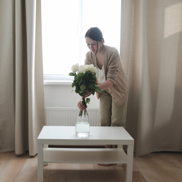 一名年轻女子在自然光下将一束新鲜美丽的玫瑰插在白色桌子上的玻璃花瓶里图片下载