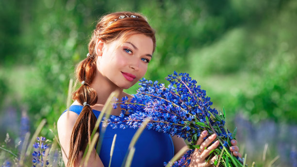 年轻美丽的姜黄色女人的肖像与蓝色的夏季羽扇花在阳光灿烂的田野。穿着蓝色衣服的红发美女在花丛中微笑。优雅的模特在自然摆姿势。图片下载