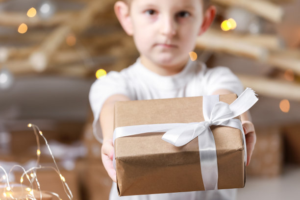 小男孩拿着圣诞背景的礼品盒。简单的礼物在牛皮纸包装系上白色的丝带在孩子的手中的t恤。元旦、母亲节、情人节图片下载