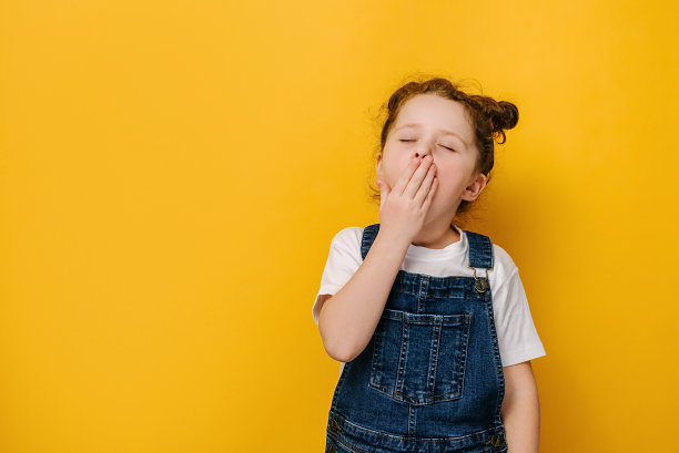 小学前班的女孩感觉累了或困了，用手捂着嘴闭着眼睛打哈欠，孤立在黄色的画室背景上用拷贝空间，脸上可爱的学前班的孩子苦不堪言。无聊的概念图片下载