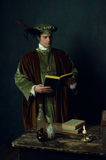 一个文艺复兴时期的人，手里拿着一本书，站在桌子后面，桌子上放着书，三个玻璃瓶和一个烛台。图片下载