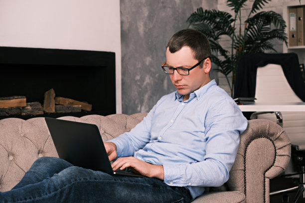 一位年轻的成功男士，穿着衬衫，戴着眼镜，坐在一间宽敞的办公室里，坐在沙发上，背靠壁炉，用笔记本电脑工作。成功与时间管理的理念图片下载