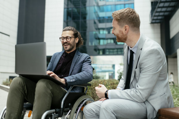 一个坐着轮椅的年轻商人和他的同事坐在外面。库存图片图片下载