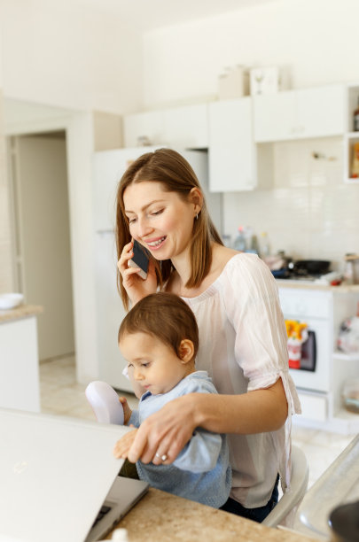 美丽的商业妈妈正在讲手机和做笔记，同时花时间与她的可爱的儿子在厨房家庭办公室。工作妈妈概念，桌上放笔记本电脑图片下载