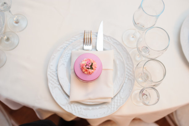 照片中的刀叉放在折叠的餐巾纸上，放在两个盘子里，旁边是一套玻璃杯，餐巾纸上有一个粉红色的圆形礼盒，上面有一朵花图片下载