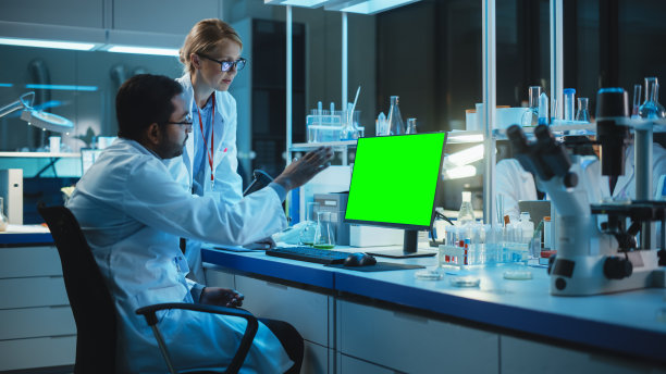 一位女研究科学家在一台绿色屏幕模拟的桌面电脑旁与生物工程师交谈。他们在现代科学实验室看电脑模板。图片下载