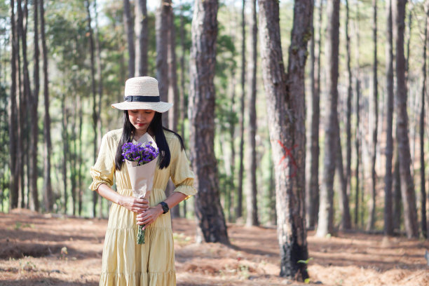 亚洲女性游客身穿黄色连衣裙，手持紫色花束，松林背景。图片下载