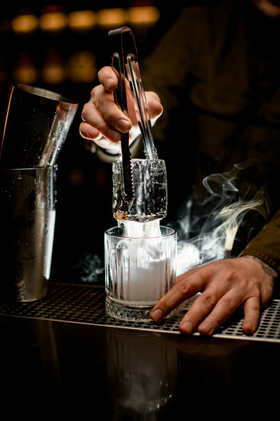 调酒师小心翼翼地用钳子夹着一大块冰块放在热气腾腾的老式玻璃杯上图片下载