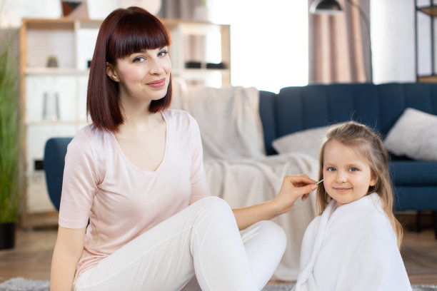 妈妈和她可爱的小宝贝女儿在洗完澡后，坐在现代化公寓室内的地板上。妈妈用棉签擦拭女儿的耳朵。近距离图片下载