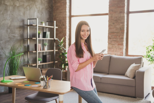 乐观的黑发女士的资料照片，拿着电话，坐在桌子上，穿粉红色的衬衫，牛仔裤，从家里学习图片下载