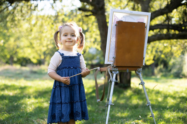 在阳光明媚的夏日公园里，可爱的小女孩身着牛仔裙，手拿画笔，站在画架旁，对着镜头摆姿势，在户外写生图片下载